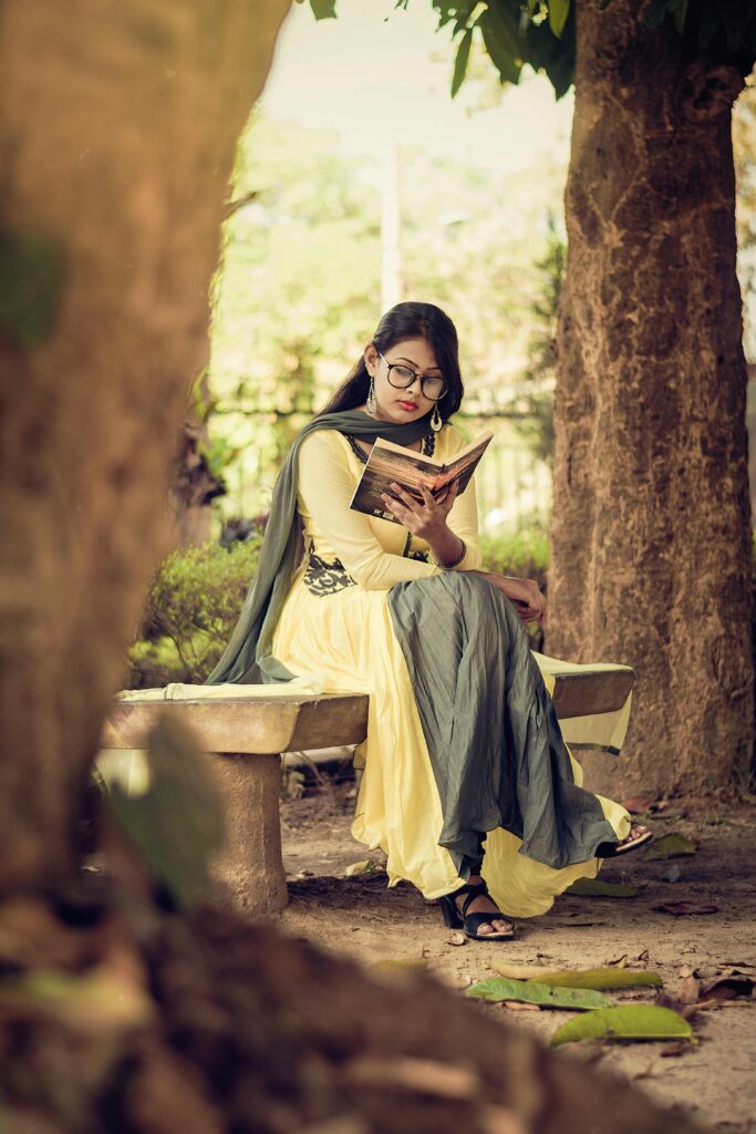 South Asian woman reading a book on a bench in a serene outdoor park setting.